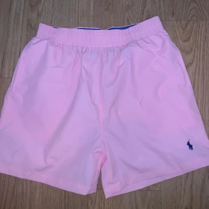 Rosa badshorts från Polo Ralph Lauren - Snygga ljusrosa badshorts från Polo Ralph Lauren med marinblå broderad logga på benet. De har elastisk midja med blå insida och meshfoder för extra komfort. Perfekta för stranden eller poolen, riktigt fräsch färg som sticker ut.