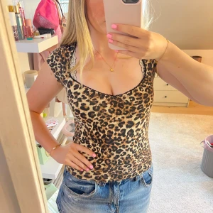 Leopardmönstrad topp med spetskant - Säljer en snygg leopardmönstrad topp med korta ärmar och djup v-ringning. Toppen har en beige och brun färgton med svarta leopardfläckar och en tunn spetskant vid halsen som ger extra detalj. Perfekt att styla med jeans för en cool look.