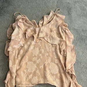 Beige blommig blus med volanger från Zara - Supersöt blus från Zara i beige med blommigt mönster och volangdetaljer längs axlar och halsringning. Blusen har en lätt glansig yta och tunna axelband, perfekt för dig som gillar romantisk stil. Materialet känns lätt och luftigt.