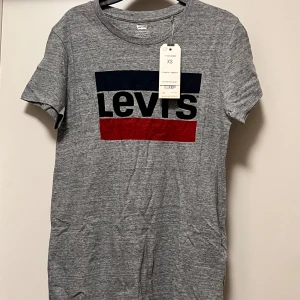 Grå Levi's t-shirt med logga XS - Snygg grå t-shirt från Levi's i mjuk bomull. Klassisk passform med rund hals och korta ärmar. Framsidan har en stor Levi's-logga i svart, blått och rött. Perfekt för dig som gillar enkel och stilren streetwear.