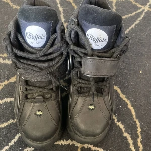 Buffalo London svarta plattformssneakers - Buffalo London sneakers i helsvart med chunky plattformssula, snörning och kardborreband över vristen. Skorna har rund tå och detaljerade sömmar, samt Buffalo-logga på plös och sidor. Materialet är skinn och syntet, perfekt för dig som gillar statement-skor.