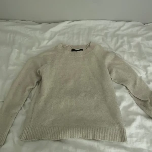 Beige stickad tröja med rund hals - Mysig beige stickad tröja med rund hals och långa ärmar. Tröjan har ribbade muddar vid ärmslut och nederkant, vilket ger en snygg och enkel look. Perfekt för att hålla sig varm under kyliga dagar.