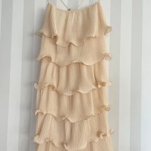 Champagnefärgad volangklänning med smala axelband - Säljer en superfin champagnefärgad klänning med smala axelband och flera lager volanger. Klänningen har plisserat tyg som ger en snygg struktur och ett luftigt intryck. Perfekt för dig som gillar romantisk och trendig stil med extra volym.