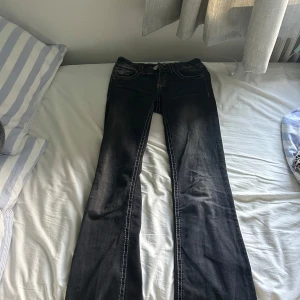 Svarta bootcut jeans med kontrastsömmar - Säljer ett par svarta jeans från Gina Tricot med tydliga vita kontrastsömmar och snygga fickdetaljer med knappar bak. Modellen är bootcut med normal passform och klassisk femficksdesign. Perfekt för dig som gillar en lite edgy stil.