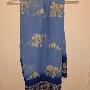 Blå sarong med elefantmönster - Snygg blå sarong/sjal med detaljerade elefantmotiv. Tunt och lätt material som ger ett luftigt intryck. Perfekt accessoar för att lyfta din outfit med ett unikt och färgstarkt mönster.