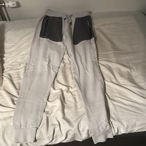 Ljusgrå mjukisbyxor från Hoodrich - Snygga ljusgrå mjukisbyxor från Hoodrich med mörkgrå detaljer vid fickorna och tryckt logga på benet. Byxorna har snörning i midjan, muddar vid bensluten och en bakficka med världskarta-tryck. Perfekta för chill eller streetwear.