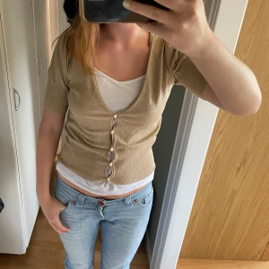 Guldglittrig beige bolero med broderi - Supersöt guldglittrig beige bolero med korta ärmar och v-ringning. Knappar framtill och broderat blommönster i brunt på ryggen. Sitter tajt och har en lite croppad passform. Perfekt för dig som vill sticka ut med lite extra shine!