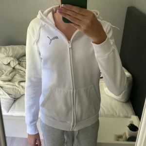 Vit hoodie från Puma med dragkedja - Säljer en fräsch vit hoodie från Puma med hel dragkedja och huva med snörning. Tröjan har ett svart Puma-logotryck med zebramönster på bröstet, två fickor fram och ribbade muddar. Perfekt för chill dagar eller när du vill ha en clean look.