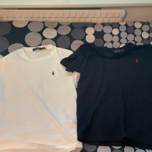 Svart och vit t-shirt Polo Ralph Lauren - Två klassiska t-shirts från Polo Ralph Lauren, en i svart med röd broderad logga och en i vit med svart broderad logga. Båda har rund hals och korta ärmar, tillverkade i mjuk bomull. Perfekt för dig som gillar stilrena och ikoniska plagg.