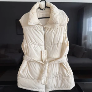 Beige dunväst från NLY Trend - Snygg beige dunväst från NLY Trend med hög krage och dragkedja framtill. Västen har ett bälte i midjan för extra stilpoäng och quiltad design som ger en cool look. Perfekt att slänga över en hoodie eller tröja när det är lite kyligt ute. Kan finnas lite foundation fläckar vid kragen men inget jag har hunnit tagit bort 