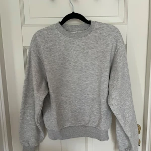 Grå sweatshirt med rund hals - En enkel och stilren grå sweatshirt med rund hals och ribbade muddar vid ärmslut och nederkant. Tröjan har en loose passform och är långärmad, perfekt för chill dagar. Materialet känns mjukt och skönt mot huden, troligtvis bomull eller polyester.