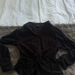 Svart skjorta med knyt från Gina Tricot - Svart skjorta från Gina Tricot i storlek XS. Skjortan har lång ärm, klassisk krage och knappar framtill. Snygg detalj med knyt i midjan som ger en cool och trendig look. Perfekt att styla med jeans eller kjol för en chic outfit.