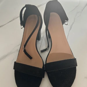 Svarta mockaklackar  - Snygga svarta klackar Skorna har en enkel rem över tårna och en justerbar rem runt ankeln med silverfärgat spänne. Tillverkade i mjuk mocka som ger en lyxig känsla och passar perfekt till dig som gillar stilrena och trendiga skor. Funkar även till 38 