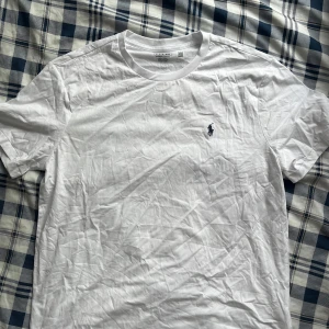 Vit t-shirt från Ralph Lauren - Vit t-shirt från Ralph Lauren med klassisk rund hals och korta ärmar. På bröstet finns den ikoniska broderade polospelaren i marinblått. T-shirten är gjord i mjuk bomull och har en clean, stilren look som funkar till allt.
