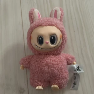 Lafufu typ? - Bilderna visar en gullig rosa mjukisdocka med stora ögon och ett lurvigt kanindräktsliknande utseende. Dockan har en nyckelring och etiketter med texten 'THE MONSTERS' och 'THE MONSTEPS'. Det finns ingen bok på bilderna.