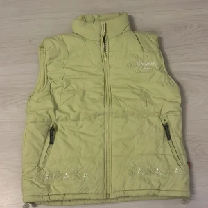 Grön dunväst med blommor från p.brand - Snygg beige dunväst från p.brand med broderade blommor längst ner fram. Västen har hög krage, dragkedja och två fickor med FIELD SPORT-detaljer. Perfekt för lager-på-lager och har en mjuk, quiltad känsla.