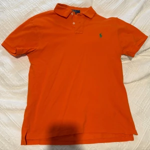 Orange polo t-shirt från Ralph Lauren - Fräsch orange polo t-shirt från Ralph Lauren i slim fit. Klassisk krage med två vita knappar och den ikoniska gröna Polo-loggan broderad på bröstet. Tillverkad i mjuk bomull som är skön mot huden. Perfekt för dig som gillar stilren och färgstark look.