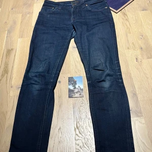 Mörkblå slim jeans från Nudie Jeans Co - Snygga mörkblå jeans från Nudie Jeans Co med klassisk femficksdesign och raka ben. Jeansen är tillverkade i kraftig denim av bomull och har subtila slitningar för en cool look. Perfekta för dig som gillar stilrena och tidlösa jeans. I storlek W27 L 30