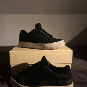 Svarta mocka sneakers från Axel Arigato - Svarta sneakers från Axel Arigato i mjuk mocka med vit platt sula. Snygg minimalistisk design utan snörning, med rund tå och diskret logga på sidan. Perfekt för dig som gillar stilrena och cleana skor med streetkänsla. Äkta!! Inte jätte bra skick! Det är bara att höra av sig om några frågor! Pris ej hugget i sten!!!!
