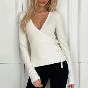 Vit omlottopp med knyt i midjan - Snygg vit omlottopp med lång ärm och v-ringad hals. Toppen har ett stilrent knyt i midjan som ger en feminin siluett. Perfekt att matcha med jeans eller kjol för en clean och fräsch look. Materialet känns mjukt och följsamt mot huden.