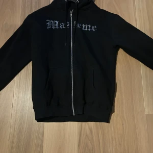 Svart rhinestone hoodie från Masteme - Svart hoodie med dragkedja från Masteme, dekorerad med rhinestone på bröstet och en stor glittrig bild av Jokern med ett rött ess på ryggen. Tröjan har huva med snörning och praktiska fickor framtill. Perfekt för dig som gillar edgy streetwear och coola detaljer.