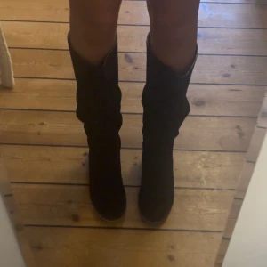 Svarta höga stövlar i mocka - Säljer ett par svarta höga stövlar med platt sula och rund tå. Stövlarna är tillverkade i mjuk mocka och har en snygg slouchy passform som ger en cool vibe. Perfekta att styla med kjol eller jeans för en trendig look.