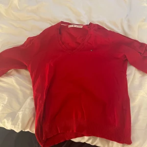 Röd v-ringad tröja Tommy Hilfiger - Snygg röd v-ringad tröja från Tommy Hilfiger med diskret logga på bröstet. Tröjan har långa ärmar och ribbade muddar vid ärmslut och nederkant. Perfekt för dig som gillar klassisk stil med en färgklick. Materialet känns mjukt och skönt mot huden.