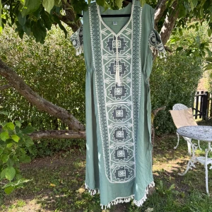 Grön långklänning med broderi - Säljer en grön långklänning med vita och gröna nyans broderade geometriska mönster framtill. Klänningen har korta ärmar med dekorativa tofsar och v-ringad hals med snörning. Materialet känns som bomull och nederkanten har vita fransar för extra boho-vibe. Storlek One Size men passar bäst kanske en XL. Myclet fint skick, jag hittar inga synliga anmärkningar eller defekter. 