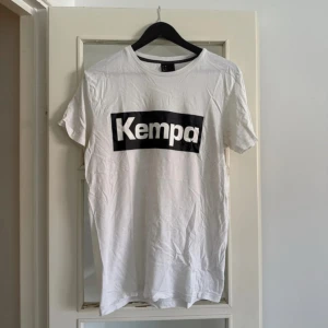 Vit Kempa t-shirt med tryck - Vit t-shirt från Kempa i storlek L med stort svart tryck över bröstet. Klassisk rund hals och korta ärmar. Mjuk och skön bomull som passar perfekt till chill eller sport. Enkel och stilren design med kontrast mellan vitt och svart.