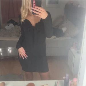 Svart kort klänning från Missguided - Säljer en svart kort klänning från Missguided med långärmade ärmar och snygg rosettdetalj framtill. Klänningen har en v-ringad urringning och sitter tight, perfekt för dig som gillar en stilren och chic look. Materialet känns mjukt och följsamt mot kroppen.