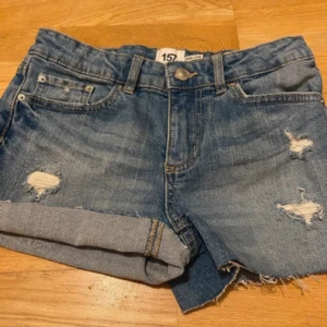 Blå jeansshorts med slitningar 157 - Snygga blå jeansshorts från 157 med coola slitningar och fransiga kanter. Klassisk femficksmodell, uppvikta ben och knappstängning fram. Perfekta för sommarens chill dagar, med en loose passform och skön denimkänsla.