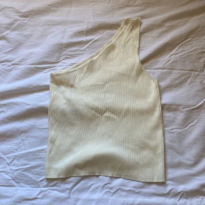Ljusbeige ribbad oneshoulder topp - Snygg ljusbeige ribbad topp med oneshoulder-design. Den är ärmlös och har en cool asymmetrisk form som ger en trendig vibe. Perfekt att styla med jeans eller kjol för en chill look. Materialet är mjukt och stretchigt för skön passform.