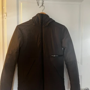 Svart vindjacka från Sail Racing XS - Svart vindjacka från Sail Racing i storlek XS. Jackan har huva, hel dragkedja framtill och två snedställda fickor. Materialet är polyester med återvunna fibrer och Primaloft-foder som håller dig varm. Snygg, stilren och perfekt för blåsiga dagar. Regn tålig, lite smutsig.