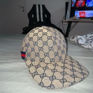 Gucci beige keps med GG-mönster - Säljer en beige Gucci keps med klassiskt GG-mönster i mörkblått. Kepsen har en snygg röd och blå rand på sidan och är tillverkad i bomull, polyester och linne med insida i mörkblått tyg. Perfekt för dig som vill ha en lyxig och trendig look.