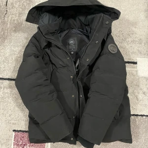 Svart dunjacka från Canada Goose - Säljer en svart dunjacka från Canada Goose med huva och snygg patch på ärmen. Jackan har dragkedja och knappar framtill, quiltad design och är långärmad. Perfekt för kalla dagar med sin rejäla och stilrena look. Passar dig som vill ha både värme och streetstyle.