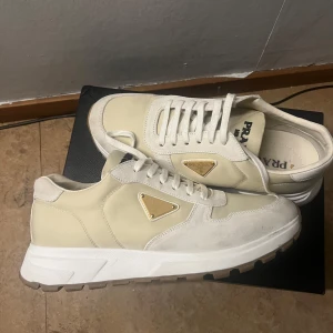 Beige sneakers från Prada - Snygga beige sneakers från Prada med vit sula och guldiga logodetaljer på sidan. Skorna har rund tå, snörning och en mix av mocka och syntetmaterial. Perfekt för dig som gillar stilrena och lyxiga sneakers med exklusiv känsla.Säljer mina Prada sneakers i beige/vitt. De är använda två gånger. Skorna är i väldigt bra skick men har lite smutsfläckar (syns på bilderna). Kvitto finns med!