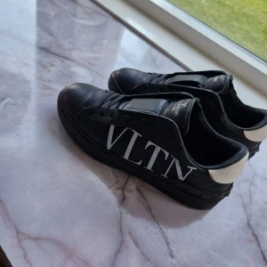 Valentino Garavani VLTN sneakers svart - Svarta sneakers från Valentino Garavani med VLTN-tryck i vitt på sidan och vit häl. Skorna har rund tå, platt sula och snörning framtill. Tillverkade i skinn med coola nitar runt sulan för extra edge. Perfekt för dig som gillar lyxiga street vibes.. Prutat och klart 1000kr. 