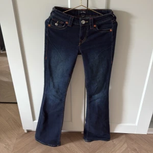True Religion bootcut jeans mörkblå - Snygga mörkblå jeans från True Religion, modell Becca Mid Rise Bootcut. Klassisk bootcut-passform med kontrastsömmar i orange och gult, samt ikoniska fickor med silverfärgade knappar och broderade detaljer. Perfekt för dig som gillar statement-jeans med coola detaljer. 