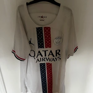 PSG x Jordan vit fotbollströja - Snygg PSG fotbollströja i vitt från Jordan med Qatar Airways tryck på bröstet. Tröjan har korta ärmar med röd, blå och vit kant, samt två vertikala ränder i blått och rött med mönster. Materialet är lätt och andas, perfekt för match eller träning.
