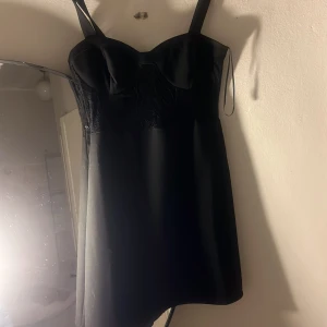 Svart kort klänning med axelband - Snygg svart kort klänning med breda axelband och markerad byst. Klänningen har en stilren, figurnära passform och slät yta med subtila detaljer upptill. Perfekt för dig som gillar minimalistisk och chic stil.