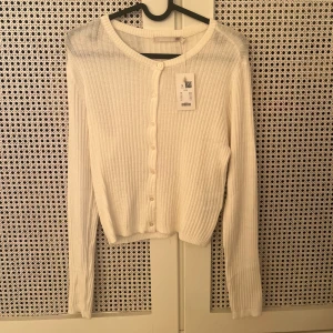 Vit ribbad kofta från lager 157 - Säljer en vit kofta från lager 157. Aldrig använd med ribbad struktur och rund hals. Koftan har långa ärmar och knappar framtill, perfekt att slänga över en topp. Materialet är mjukt och stretchigt, vilket gör den skön att bära. Passar dig som gillar enkel och clean stil.