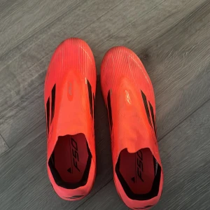 Adidas F50 röda fotbollsskor - Säljer ett par Adidas F50 fotbollsskor i en riktigt snygg röd färg med svarta detaljer och klassiska tre ränder på sidan. Skorna har en strömlinjeformad design utan snörning och är gjorda i syntetmaterial för lätthet och snabbhet på planen. Perfekt för dig som vill sticka ut på matchen.