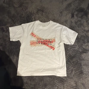 Vit t-shirt Maison Margiela med tryck - Säljer en clean vit t-shirt från Maison Margiela med unikt grafiskt tryck i rött och svart på framsidan. T-shirten har klassisk passform, rund hals och korta ärmar. Materialet är mjuk bomull som känns skönt mot huden. Perfekt för dig som gillar statement-plagg.