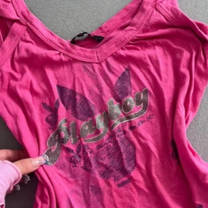 Playboy tank top - Cool rosa topp från Playboy med glittrigt tryck framtill och ikonisk kaninlogga. T-shirten har v-ringning och är gjord i mjuk bomull, perfekt för dig som gillar statement-plagg och vill sticka ut lite extra. Medium men passar även S🩷