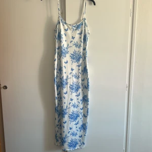 Vit långklänning med blått blommönster - Säljer en superfin långklänning med smala axelband och rundad hals. Klänningen är vit med blått blommigt mönster och fjärilar, vilket ger en fräsch och somrig vibe. Materialet känns mjukt och lätt, perfekt för varma dagar. Passformen är normal och klänningen är ärmlös.
