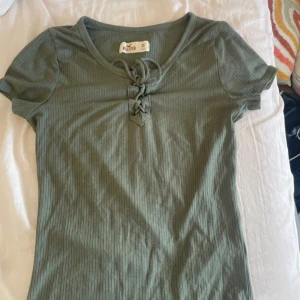 Grön ribbad topp med snörning Hollister - Snygg grön ribbad topp från Hollister i storlek XS. Modellen har korta ärmar och cool snörning framtill vid halsen. Materialet är mjukt och stretchigt, perfekt för en chill och trendig look. Passformen är normal och toppen har en lätt v-ringad hals.