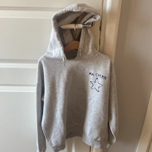 Grå hoodie med stjärna från Gina Tricot - Säljer en ljusgrå hoodie från Gina Tricot med coolt tryck 'MA CHÉRIE' och en stjärna både fram och bak. Tröjan har huva med snörning och ribbade muddar. Perfekt för chill dagar och enkel att matcha med jeans eller leggings.