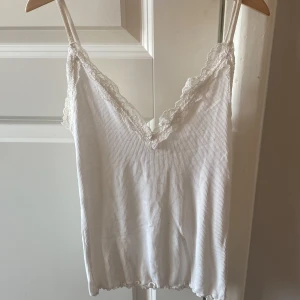 Vit ribbad linnetopp med spets New yorker - Supersöt vit linnetopp från new yorker i ribbad bomull med tunna justerbara axelband. V-ringad med spetsdetaljer längs halsringningen som ger en romantisk vibe. Perfekt att styla med jeans eller kjol för en chill look.