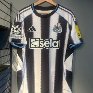 Newcastle United Isak 14 matchtröja - Snygg Newcastle United fotbollströja med Isak 14 på ryggen. Adidas-logga på bröstet, klubbmärke och sponsortryck. Svart och vit randigt mönster med blå detaljer vid krage och ärmslut. Kortärmad och tillverkad i lätt funktionsmaterial. Helt ny med tagg en replika 