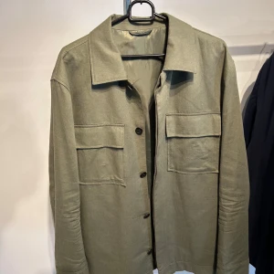 Olivgrön overshirt med fickor - Snygg olivgrön overshirt med klassisk krage och stora bröstfickor med lock. Jackan har knappar framtill och tryckknappar vid ärmsluten. Perfekt att slänga på över en hoodie eller t-shirt för en chill look. Materialet känns som bomull och har en mjuk finish.